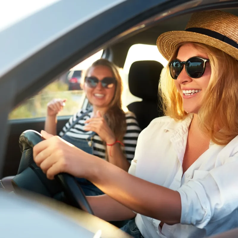 Two women in sunglasses smiling in the front seats of a car on a sunny day, the driver wearing a straw hat with her hand on the steering wheel.