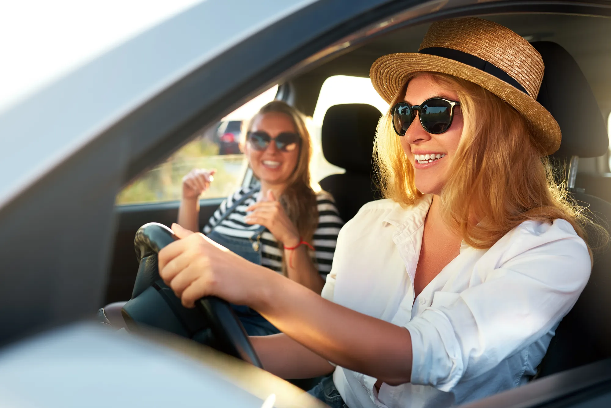 Two women in sunglasses smiling in the front seats of a car on a sunny day, the driver wearing a straw hat with her hand on the steering wheel.
