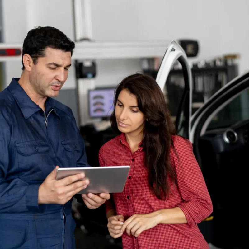 A mechanic in blue coveralls shows a tablet to a woman customer beside an open car door inside an auto repair shop.