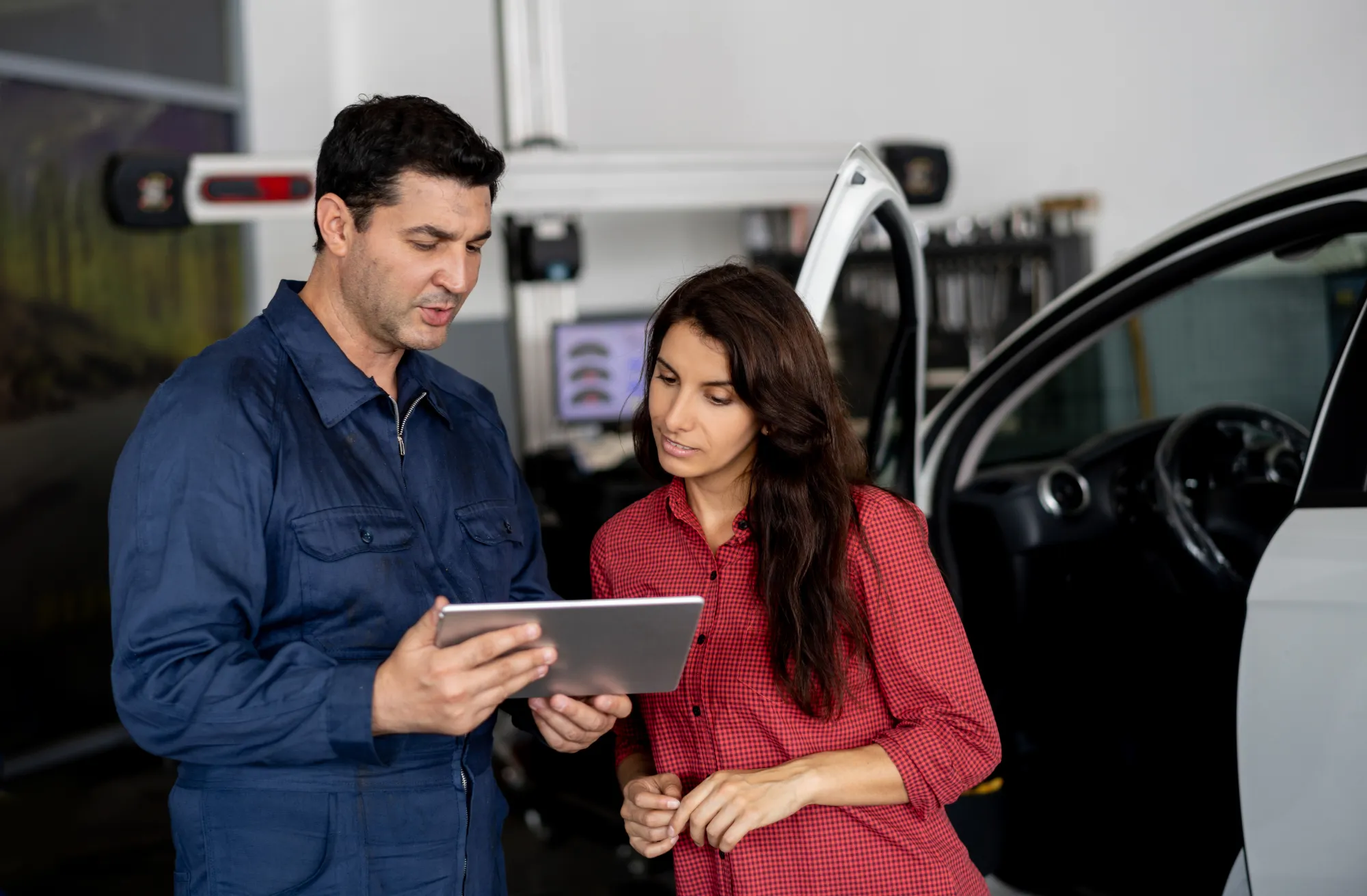 A mechanic in blue coveralls shows a tablet to a woman customer beside an open car door inside an auto repair shop.